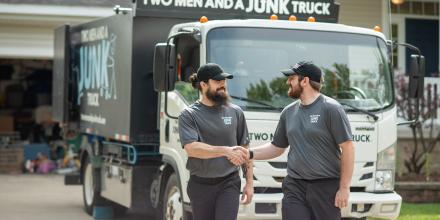 Two junk haulers shake hands in front of a TWO MEN AND A JUNK TRUCK